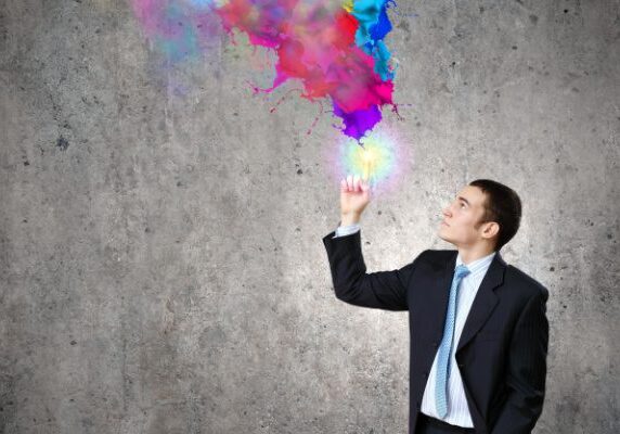 man in suit in front of concrete wall holding finger up which is alight and emitting splattered paint in many colors above it