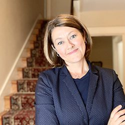 Amanda Stanley ACC headshot photo, blond smiling woman in dark top standing in a hallway in front of stairs