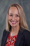 Anita Bowness headshot photo, smiling woman with brown hair wearing black blazer and red top in front of gray background