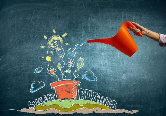 person holding watering can in front of chalk board with Success and Business written around a patch of grass with a pot on it and growing flowers money and lightbulb