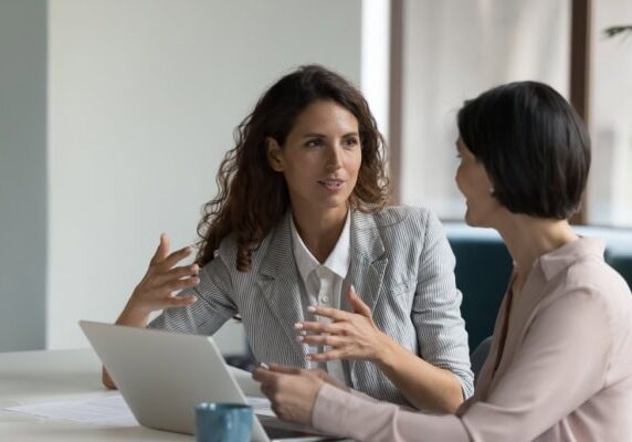 coach speaking to client and showing them something on a laptop while client speaks using her hands