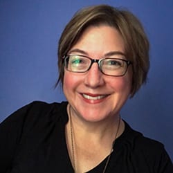 Beth Buelow PCC headshot photo, smiling woman with brown hair wearing glasses and a dark top in front of a blue background