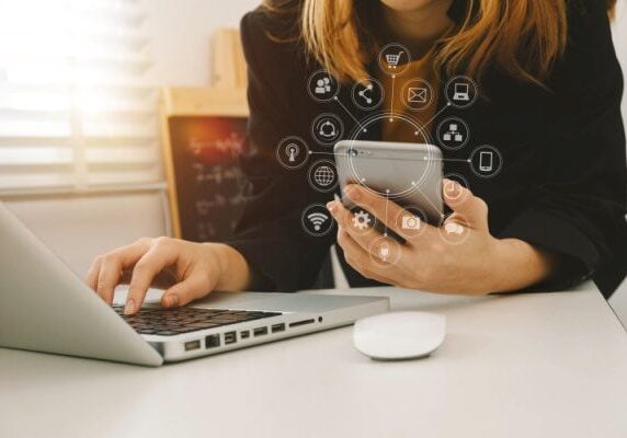 woman working on a laptop and holding a cellphone with an overlay of many icons around the cellphone