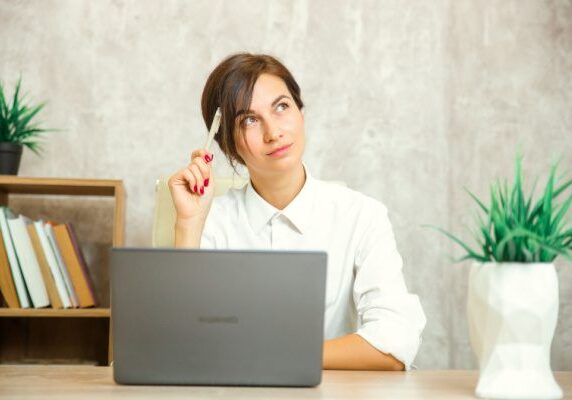 woman thinking with pen resting on head sitting in front of a laptop at a table in a home office