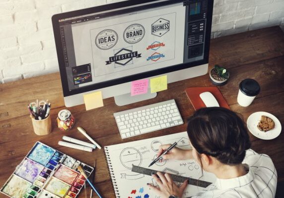 female doing grahic design at a laptop with a notebook and other art supplies to create logos