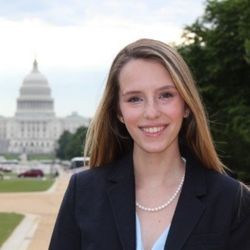 Tory Hyde headshot photo, smiling woman in black blazer standing outside of a white building near a tree
