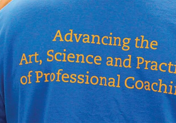 The back of a blue T-shirt worn by a person, displaying the phrase "Advancing the Art, Science, and Practice of Professional Coaching" in yellow text.