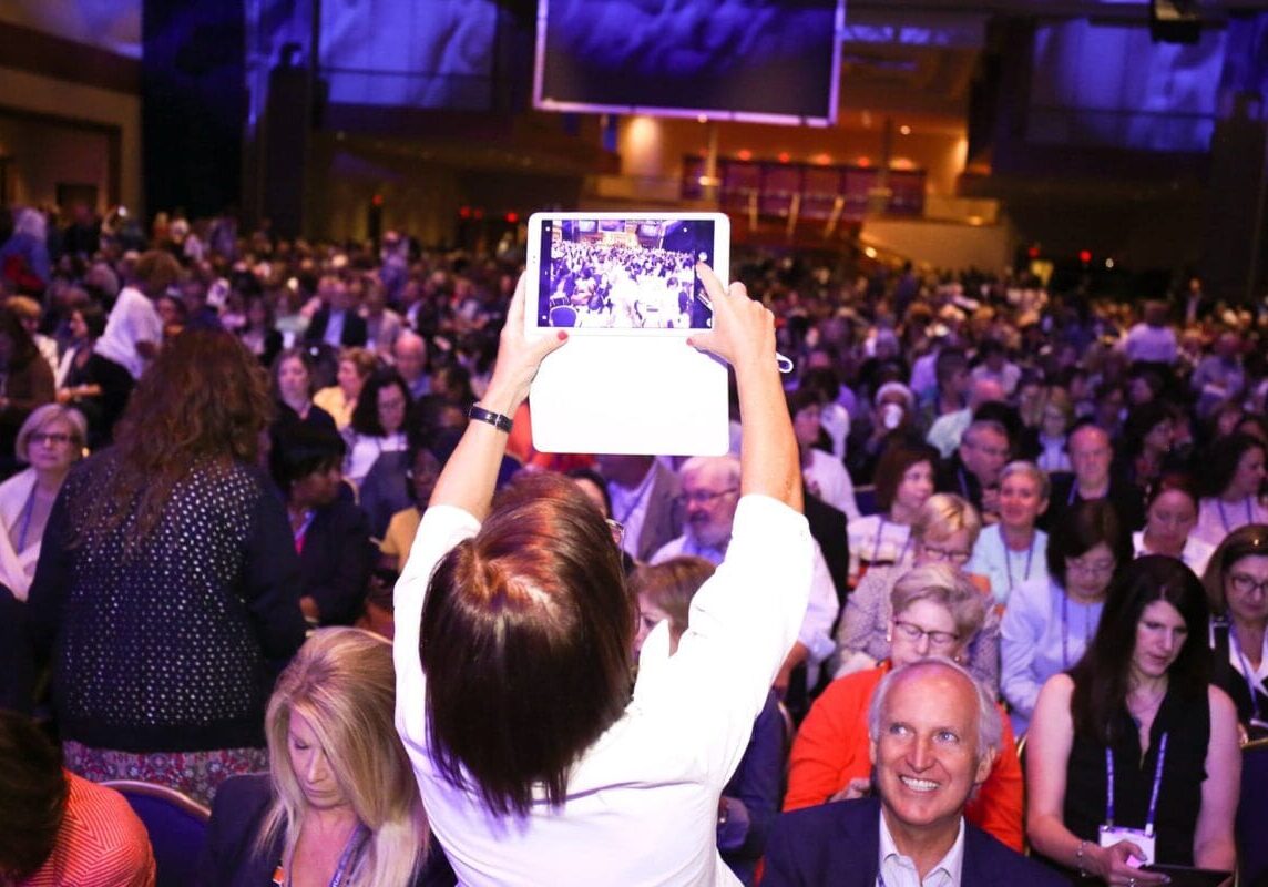 A woman in the foreground stands up and takes a photo of a diverse and inclusive crowd using her digital tablet, capturing the vibrancy of the community.