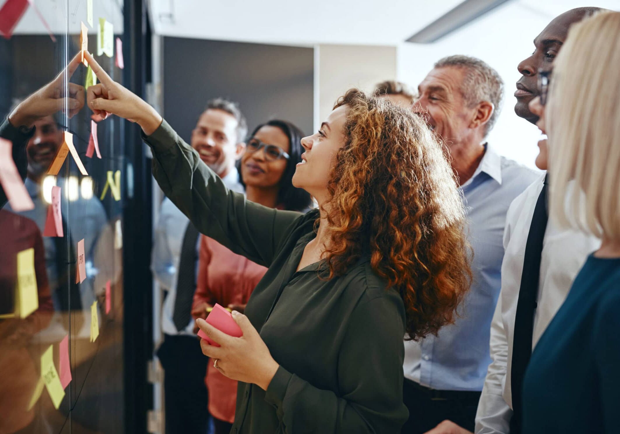 Group of professionals having a collaborative brainstorming session together, pointing at sticky notes on a glass wall, highlighting teamwork.