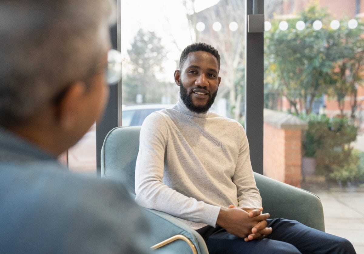 In a bright office with an urban street view, a smiling Black man in beige sweater sits in modern chair having a conversation with an individual we can only see the back of.