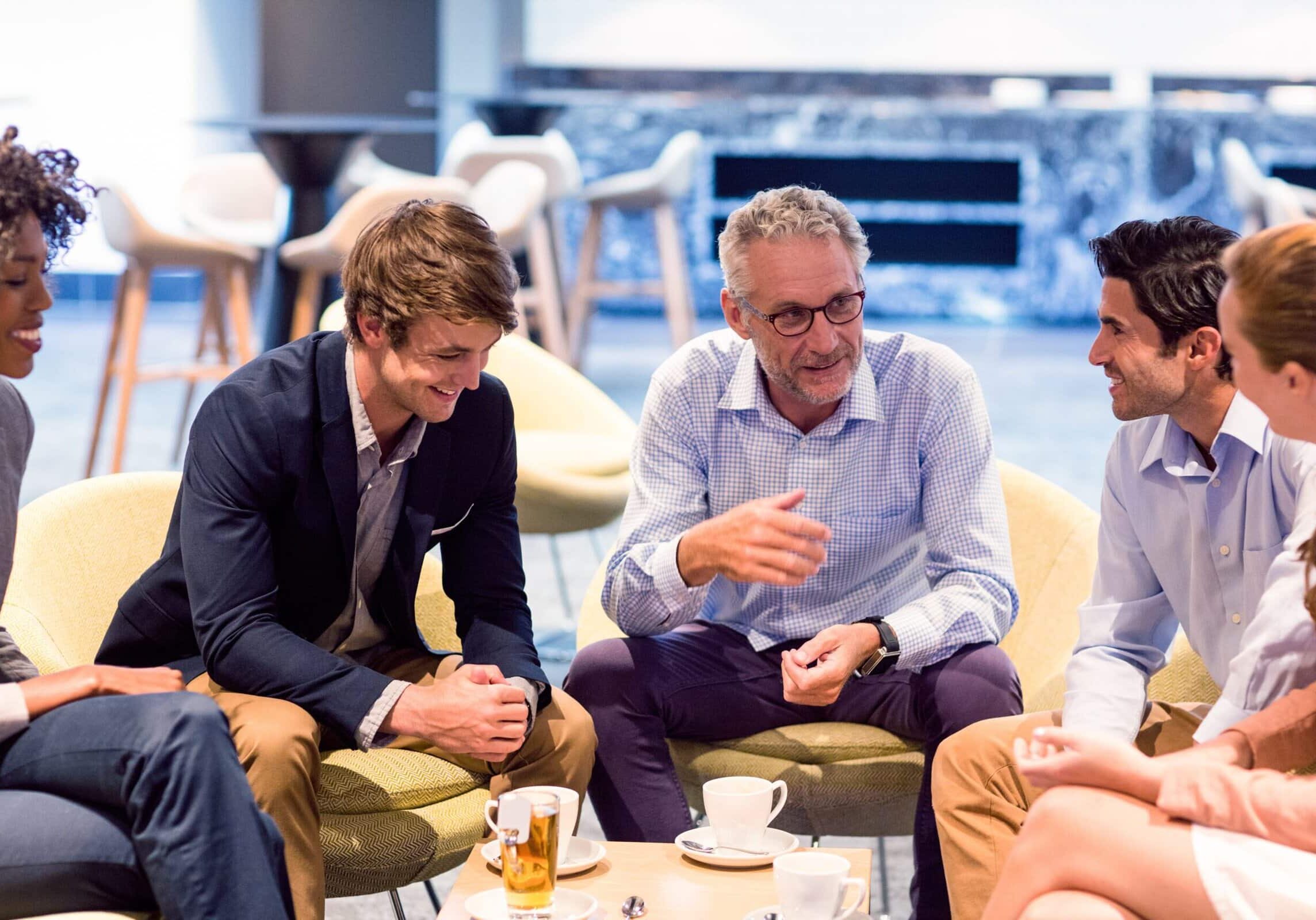 A group of five professionals seated in a lounge, discussing 168极速一分钟赛车官方开奖历史记录赛果直播预测 coaching and smiling over coffee, fostering teamwork and collaboration.
