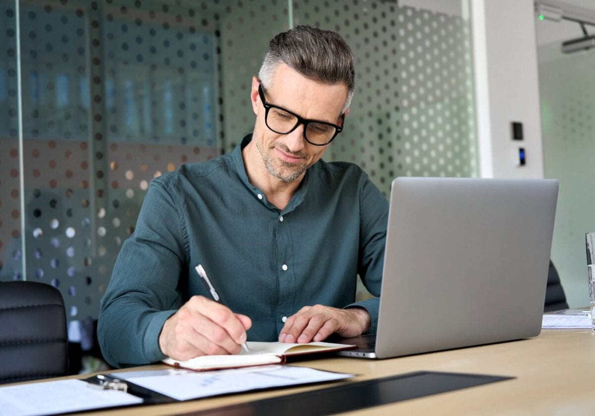 A focused, middle-aged businessman in glasses working at his desk, writing in a notebook next to a laptop.