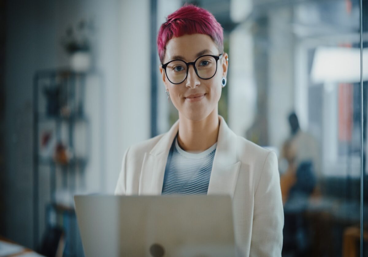   Portrait of a young, Caucasian woman with short pink hair standing, holding a laptop, looking at camera, smiling .