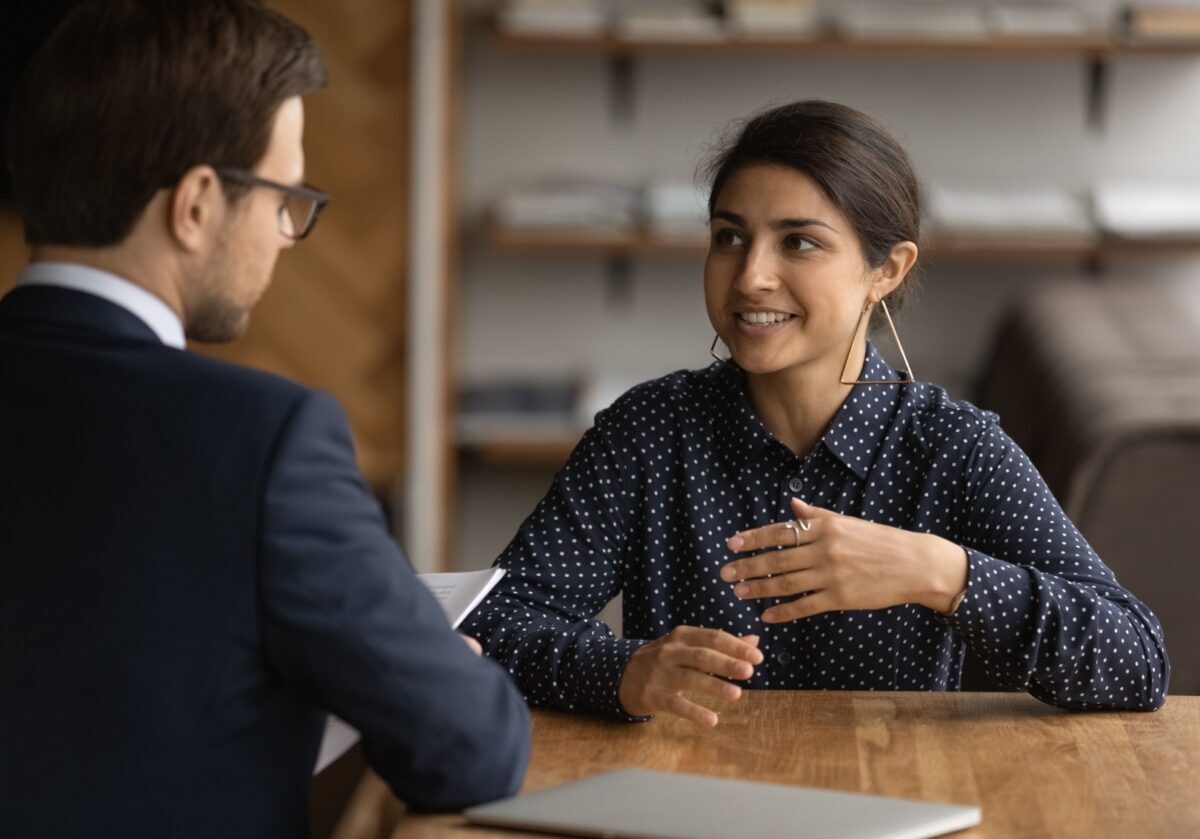 Smiling Indian woman in blue polka dots speaking with a male colleague at a table.
