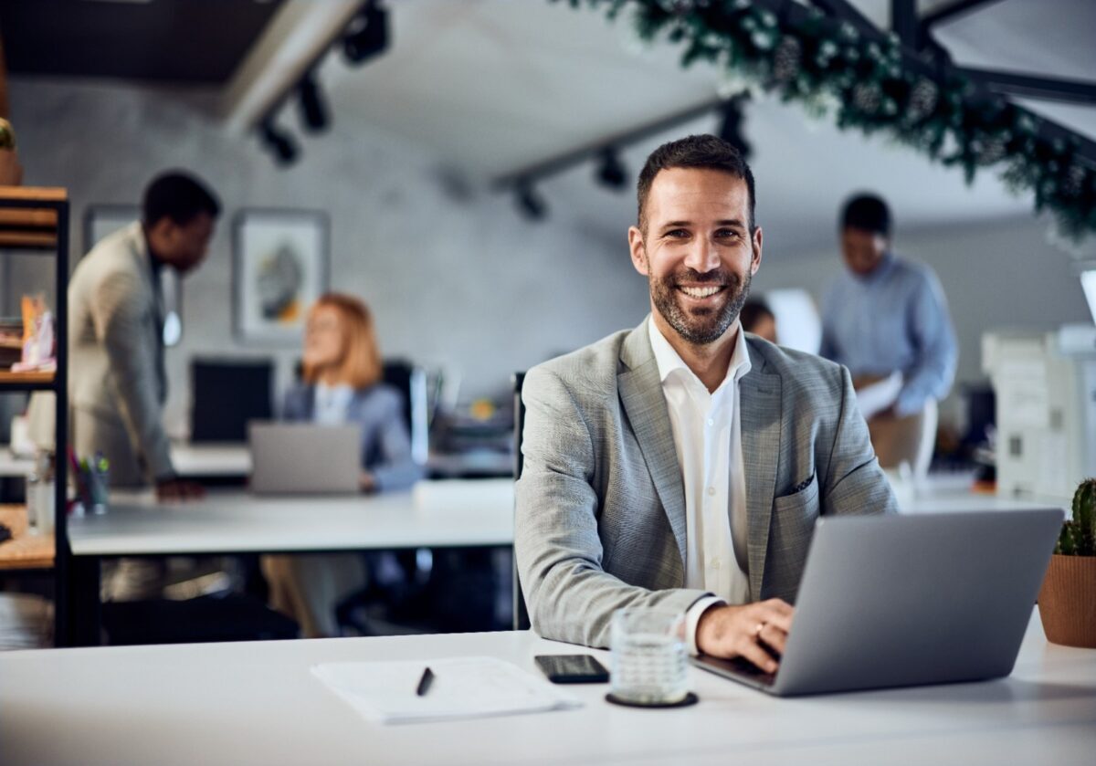 Portrait of a smiling man, working at the office, over the laptop while his colleagues are in the background.