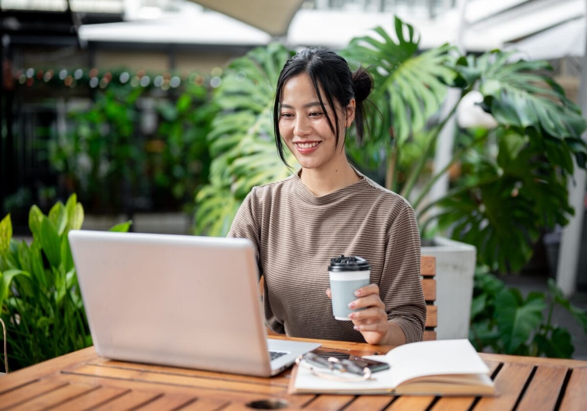 A smiling young woman sitting outdoors with a laptop, holding a coffee cup, surrounded by lush green plants.