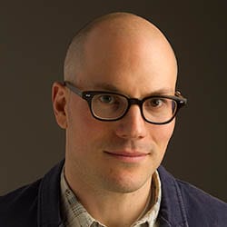 John Andrew Williams MCC headshot photo, bald man smiling with glasses and a dark blazer