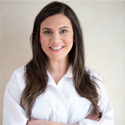 Lori Kennedy headshot photo, woman with long dark hair in white top crossing arms over chest smiling against light colored wall