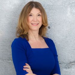 Sandra Kramer Kowalzik MBA headshot photo, woman standing against concrete wall in blue dress and long light colored hair smiling