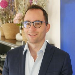 Matthias Laeubil PCC headshot photo, man with dark hair and glasses wearing a dark blazer and white collared shirt smiling in an office