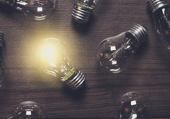 one lit lightbulb on a dark wood table with other unlit lightbulbs