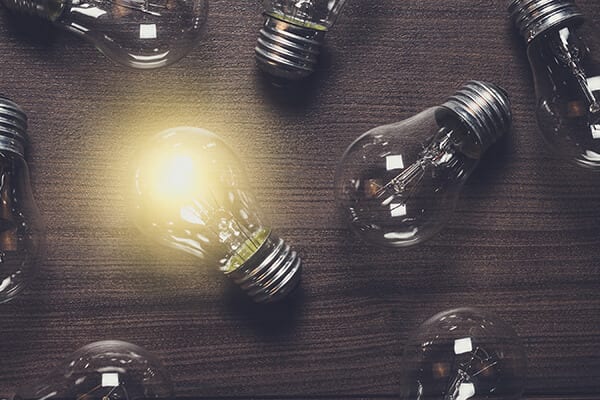 one lit lightbulb on a dark wood table with other unlit lightbulbs