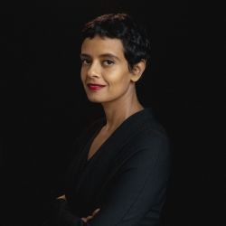 Reem Mansour PHD headshot photo, woman with short dark hair smiling with arms crossed against black background