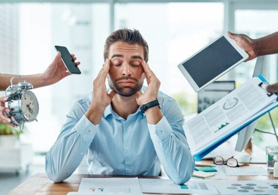 man looking stressed with hands on face and eyes closed, surrounded by various objects signifying demands and responsibilities
