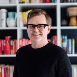Joe Oliver PHD headshot photo, man with short brown hair and black glasses wearing a black shirt standing in front of a bookshelf