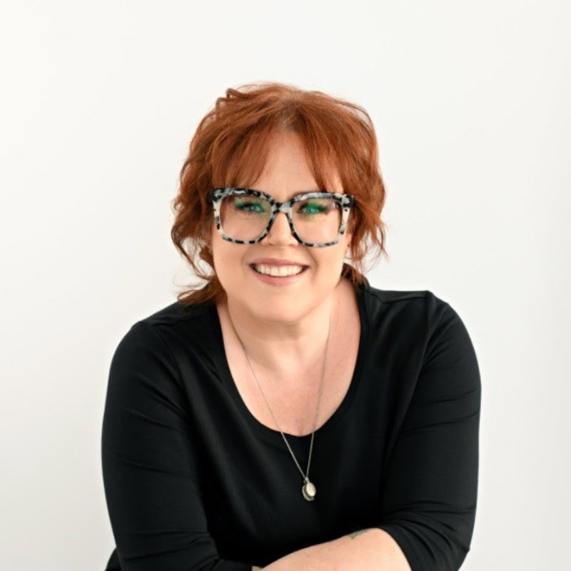 Headshot of Kels Davidson, woman wearing glasses and a black shirt with red hair smiling in front of a white background