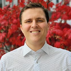 Patrick Parker PCC headshot photo, smiling man with short brown hair and light button up shirt standing in front of a red tree