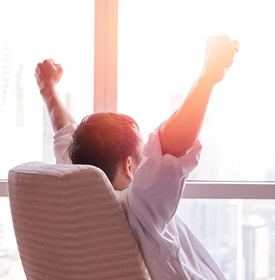 seated male in front of a window raising arms up in victory