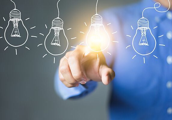 Person touching an overlayed drawing of a lightblub with it emmitting light amongst other unlit lightbulb drawings