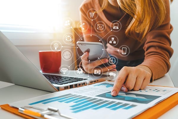 woman looking at data on a printed sheet and holding a cellphone with many connected dots and icons around it at a table with a laptop and coffee cup