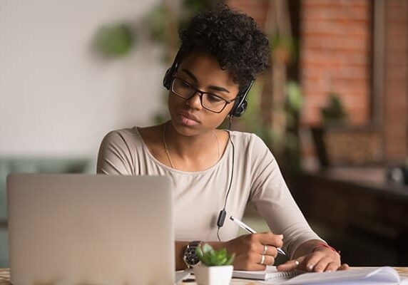 woman listening to audio from a laptop and taking notes