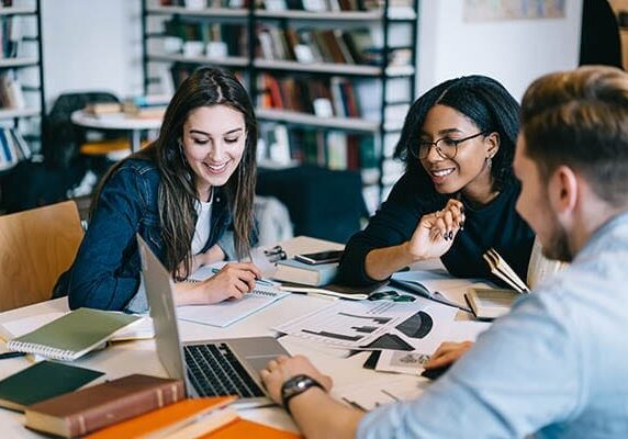 three people collaborating at a table looking at a laptop with books and papers scattered