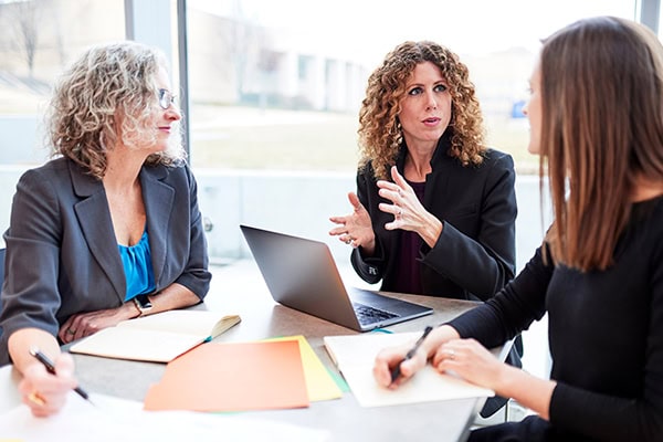 three woman talking while seated at a table with notebooks, papers, and a laptop