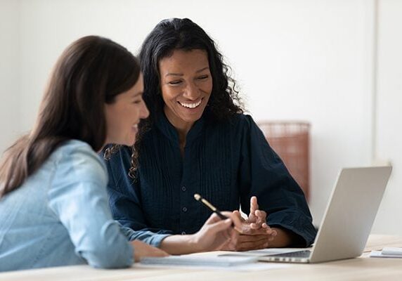 two women sitting at a laptop taking notes and engaging in conversation