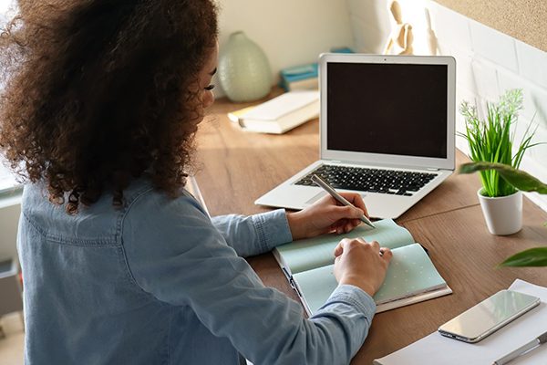 woman writing in a notebook at a desk with phone laptop plant and other papers