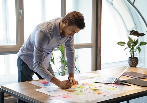 man working at a table with papers and sticky notes scattered as well as a laptop lamp and plant