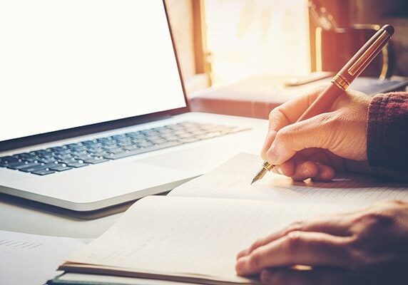 person writing in a notebook in front of a laptop on a table