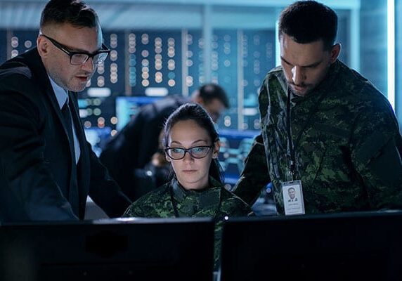 a business professional and a military person looking over the shoulder of another military service member's shoulder at her two monitors in discussion