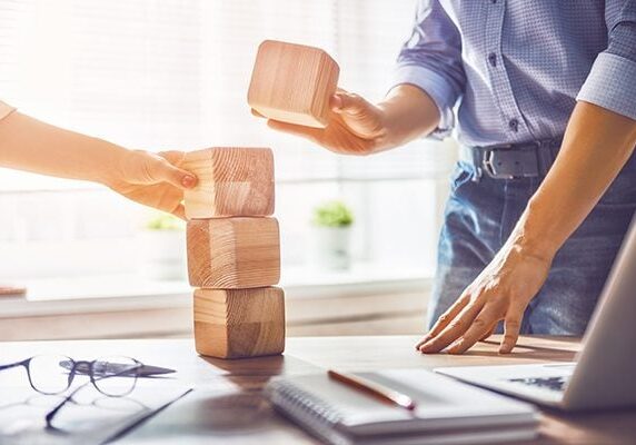 two people stacking large cube blocks on a desk with a laptop notebook papers pencil pen and glasses