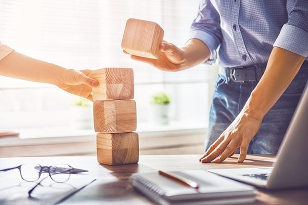 two people stacking large cube blocks on a desk with a laptop notebook papers pencil pen and glasses