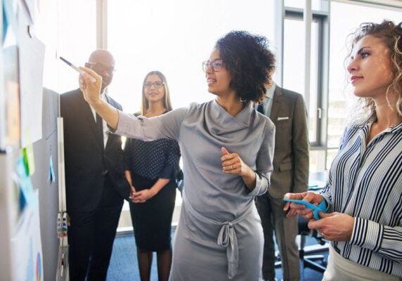 coach pointing to something on a whiteboard with four others listening, all standing in a conference room