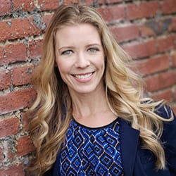 Stacey Hagen headshot photo, smiling blond haired woman with dark blue top and blazer in front of a brick wall