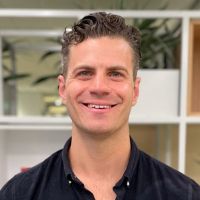 Andrew White headshot photo, man with dark brown short curly hair and dark top smiling blurred office background