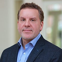 Dr William Sparks headshot photo, brown haired man smiling wearing dark blue blazer light blue shirt against blurred background